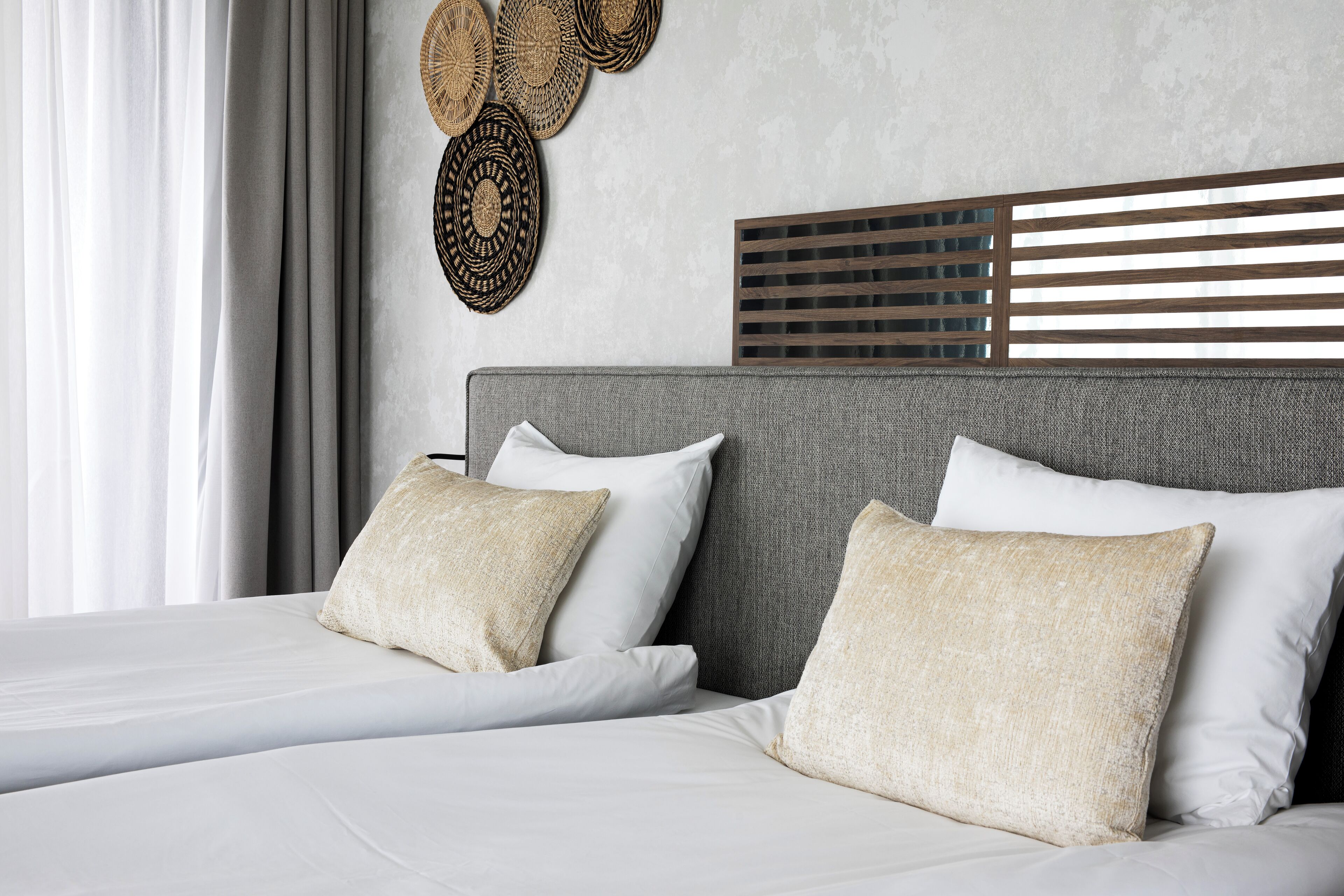 Una cama bien tendida con sábanas blancas y dos almohadas color beige, un cabecero gris y decoración mural tejida.