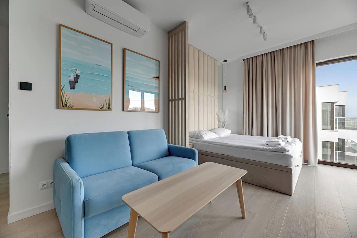 Una habitación de hotel moderna con un sofá azul, una mesa de madera, una cama con colchón y un ventanal con cortinas.