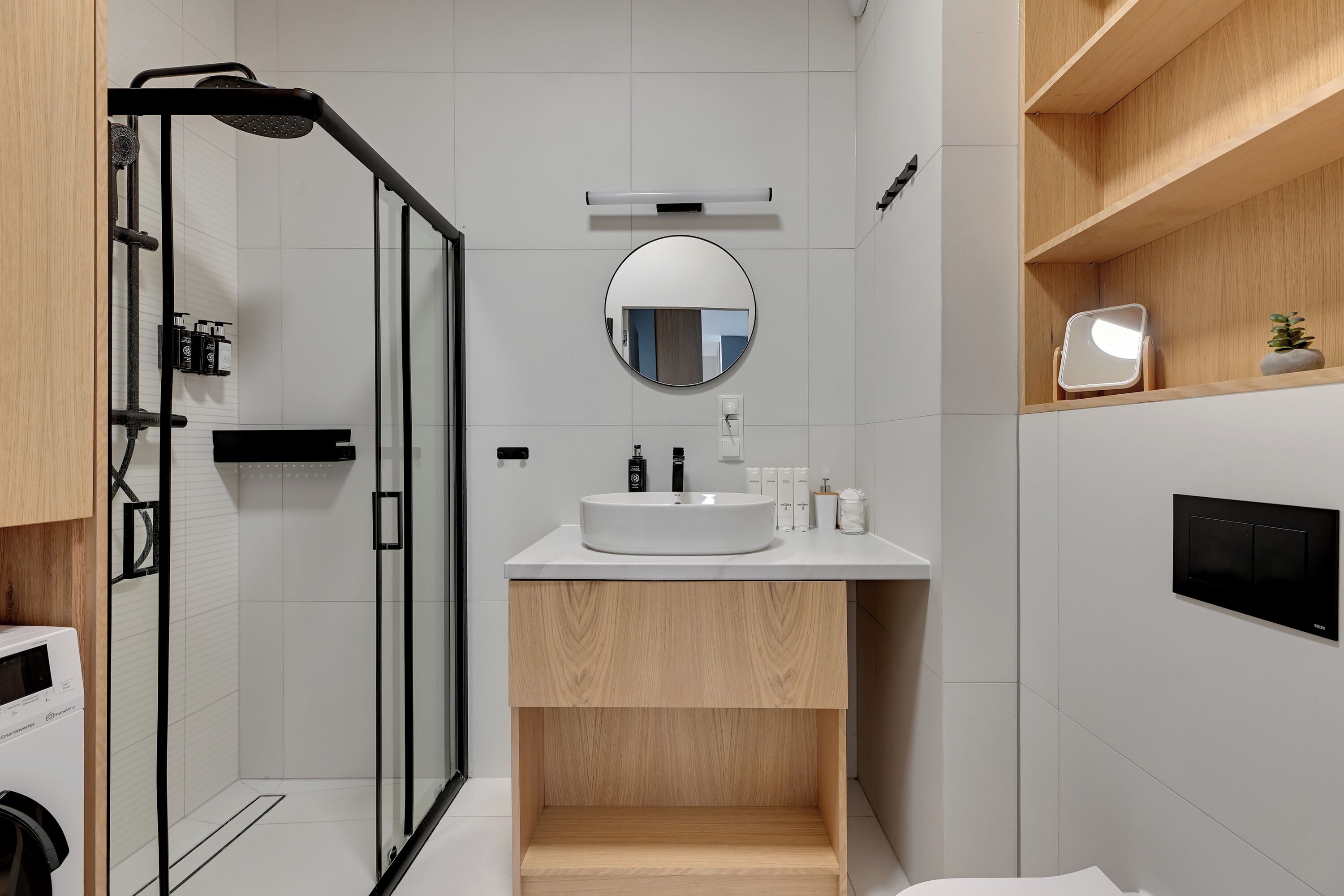 Un baño moderno con ducha envuelta en vidrio, un espejo redondo, un lavamanos blanco y estantes de madera.