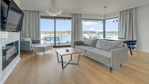 Een moderne woonkamer met een bank, salontafel en televisie, grote ramen met uitzicht op zee, en een eethoek met blauwe stoelen.