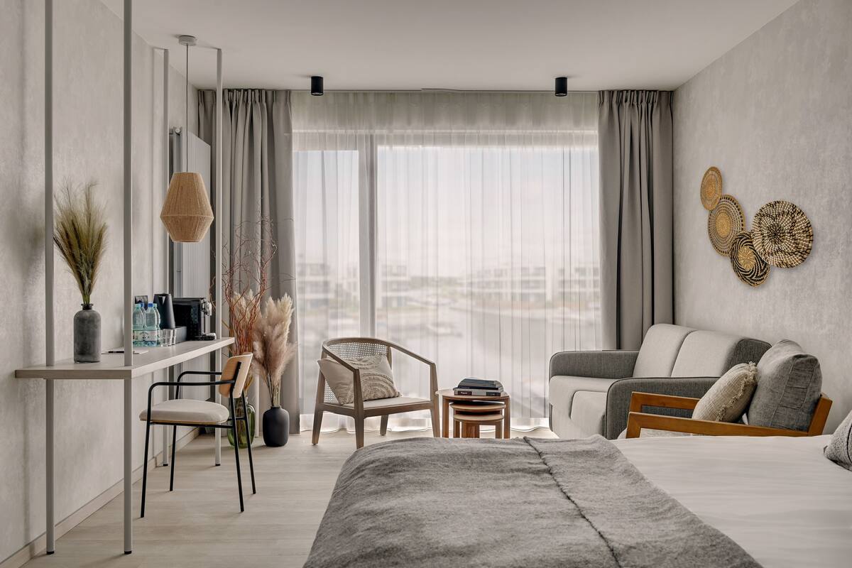 Una habitación de hotel moderna con una cama grande, un escritorio, una silla, un sofá y vistas a la ciudad a través de amplias ventanas.