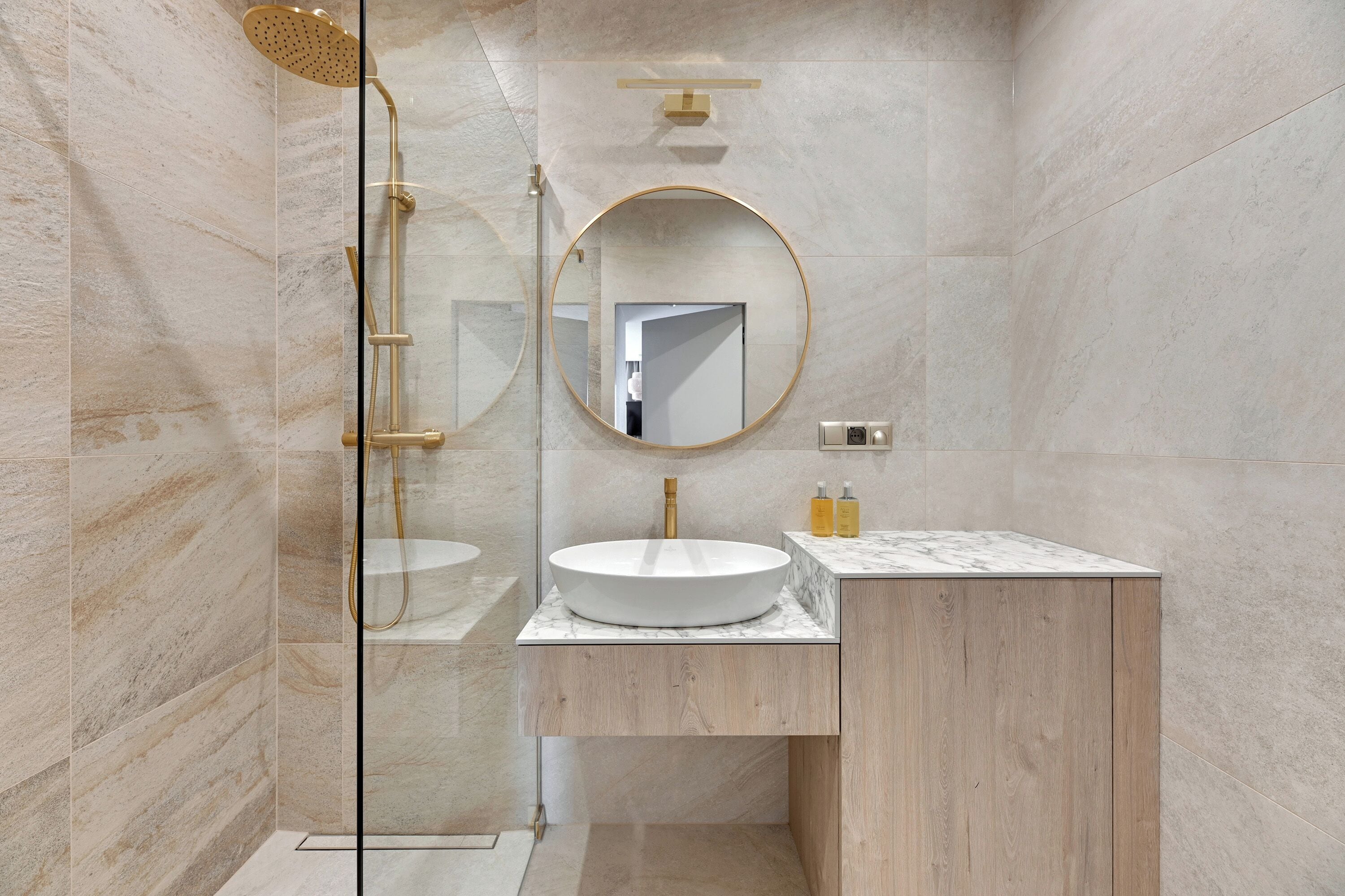 A modern bathroom with a glass-enclosed shower, a round mirror, a white sink, and wooden cabinetry.