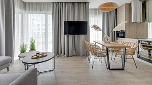 Een moderne woonkamer met een flatscreen televisie, een eethoek met rieten stoelen en een groot raam met dunne gordijnen.