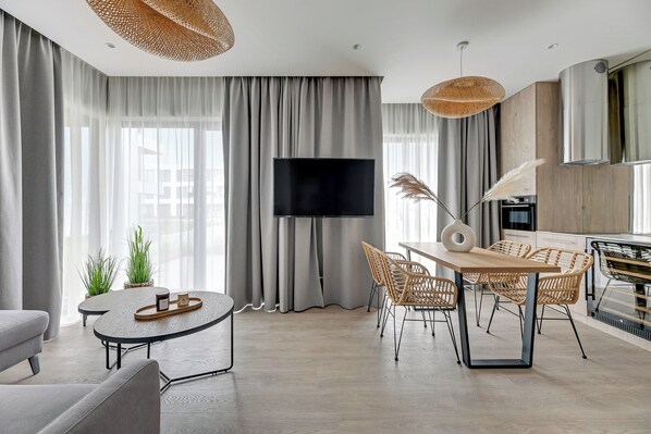 Ein modernes Wohnzimmer mit Flachbildfernseher, einem Essbereich mit Korbstühlen und einem großen Fenster mit durchsichtigen Vorhängen.