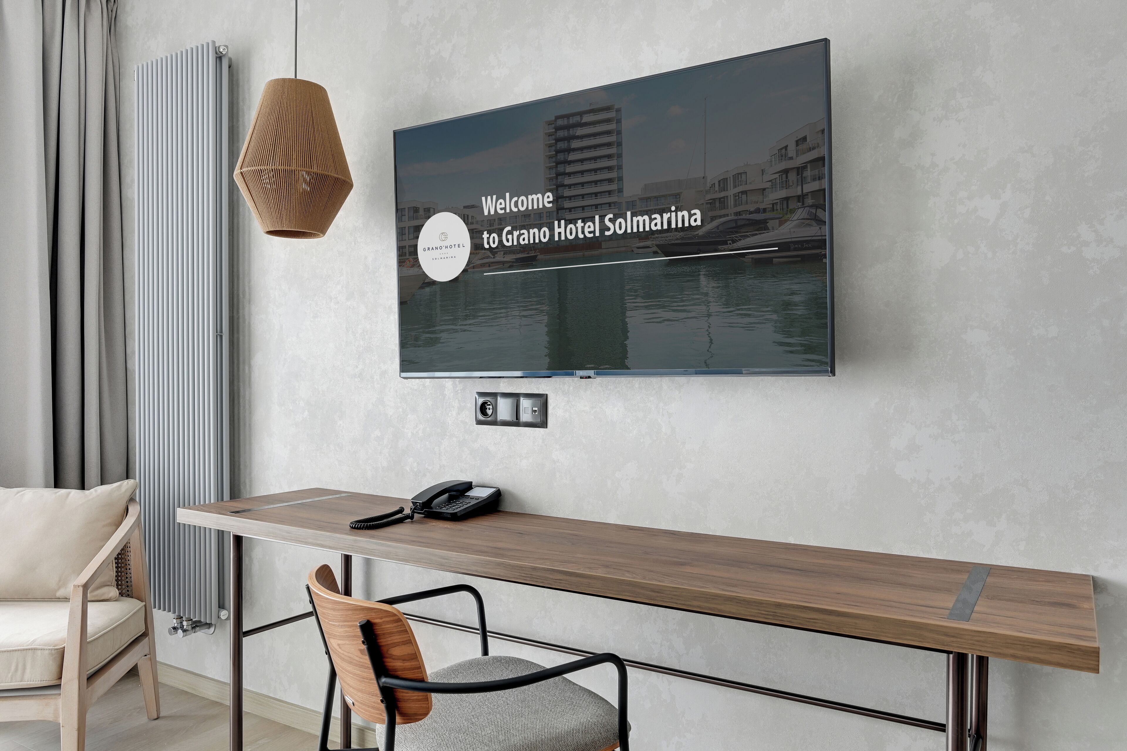 Ein modernes Hotelzimmer mit einem großen Flachbildfernseher an der Wand, der eine Willkommensbotschaft zeigt, einem Holztisch mit Telefon, einem Sessel und einer Heizung.