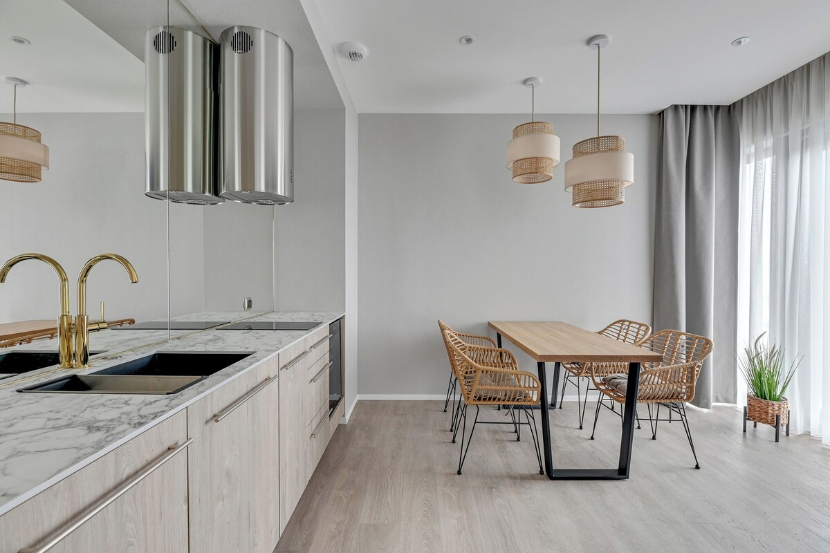 Une cuisine moderne dotée d’un plan de travail en marbre, d’une hotte en acier inoxydable et d’un coin repas avec des chaises en osier et une table en bois.