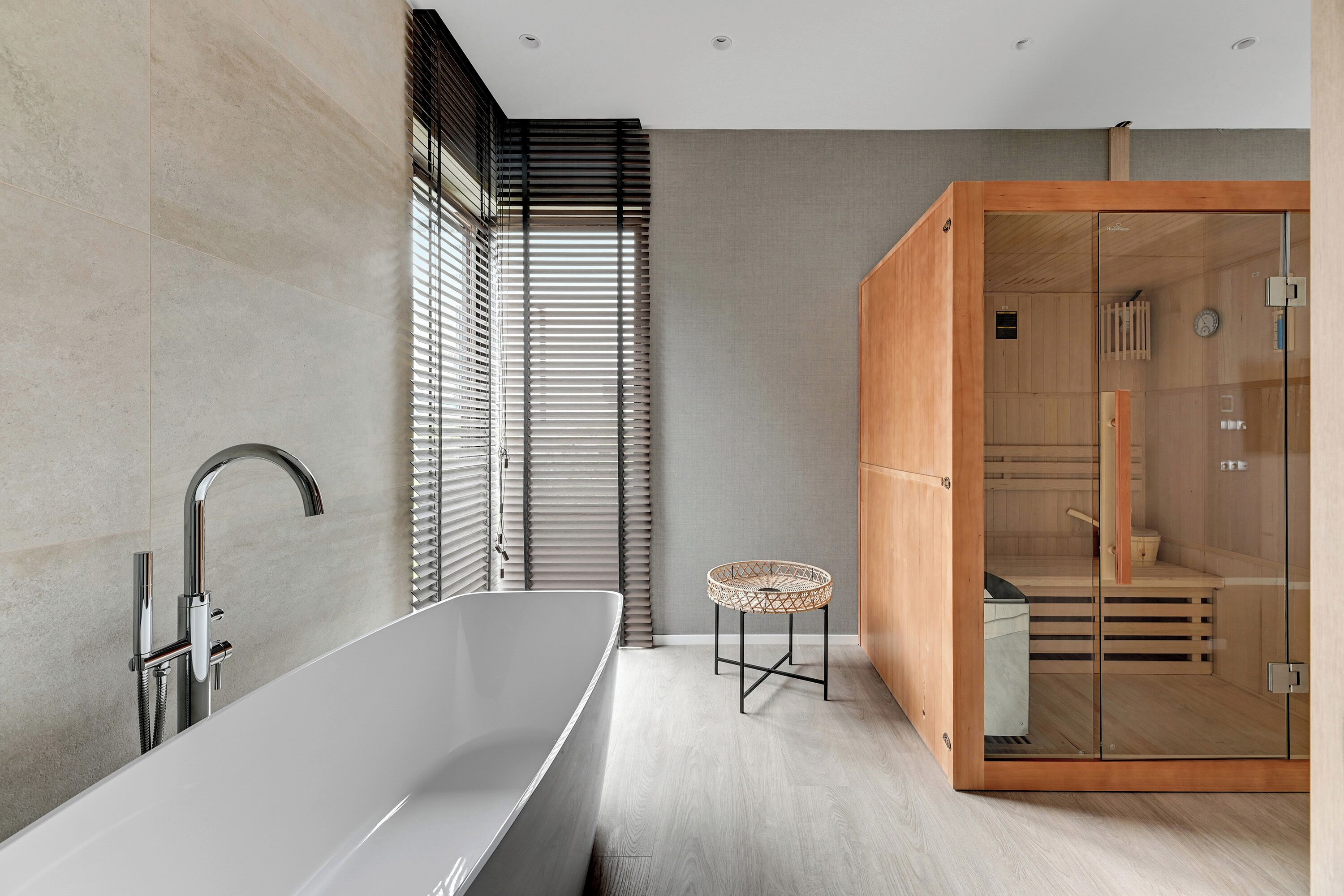 A modern bathroom with a freestanding bathtub, a glass-enclosed sauna, and a small wicker stool.