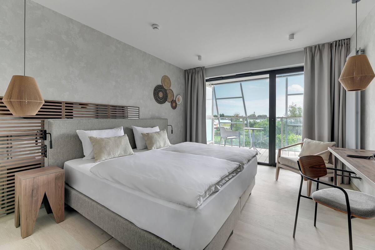 Un dormitorio moderno con una cama grande, una mesita de noche de madera, un escritorio con silla, y vistas al exterior a través de un ventanal amplio.