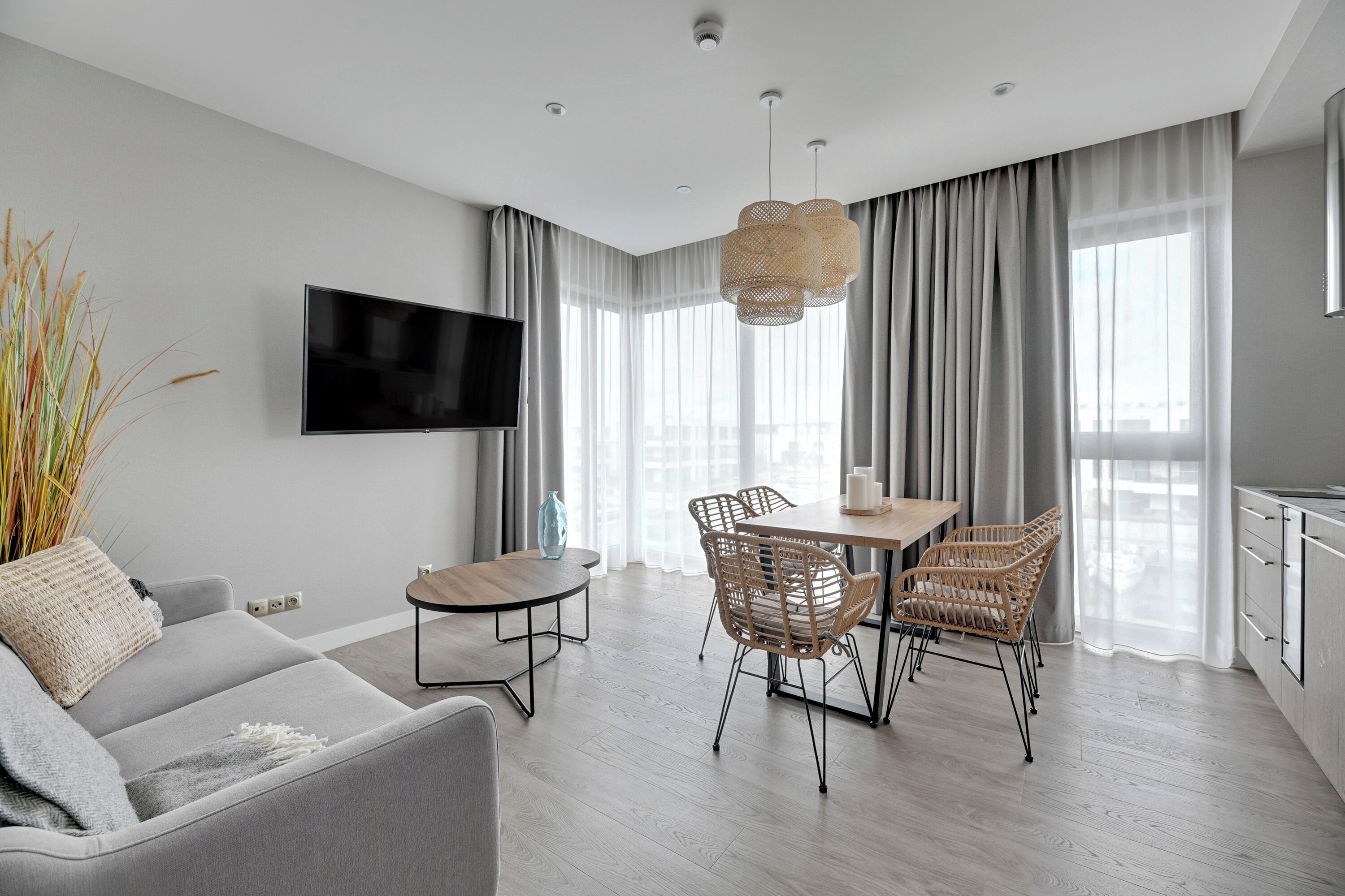 Una sala de estar moderna con un sofá gris, una mesa de comedor de madera con sillas de mimbre, un televisor de pantalla plana montado en la pared y un amplio ventanal con cortinas translúcidas.