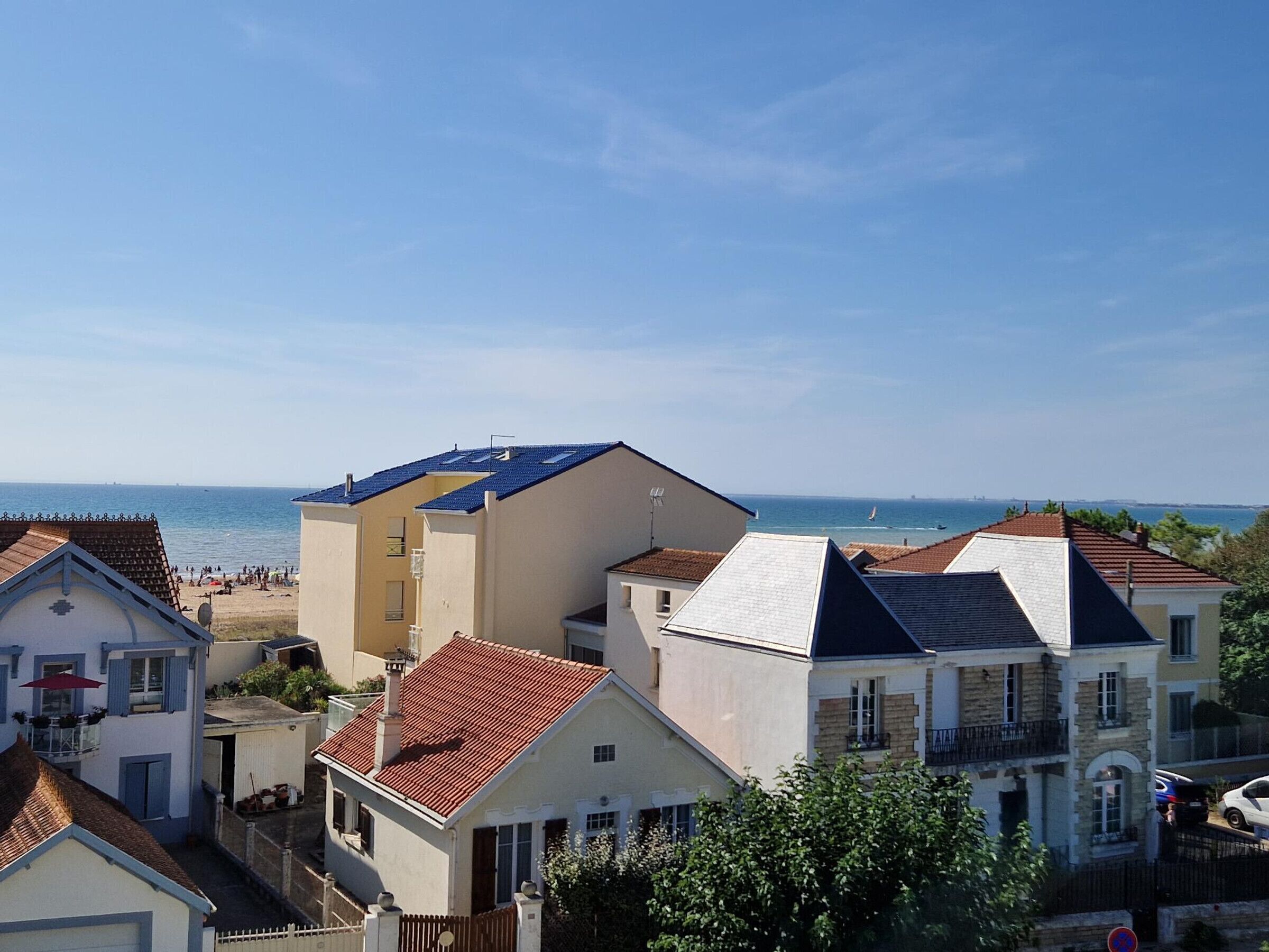 Un quartier résidentiel en bord de mer, avec des maisons aux toits de tuiles rouges et aux murs blancs, qui surplombe une plage où l’on peut voir des gens et des voiliers.
