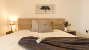 Ein ordentlich bezogenes Bett mit einer braunen Decke, einer Nachttischlampe, einem gerahmten Baum-Silhouettenbild und einem kleinen Topfpflanz auf dem Nachttisch.