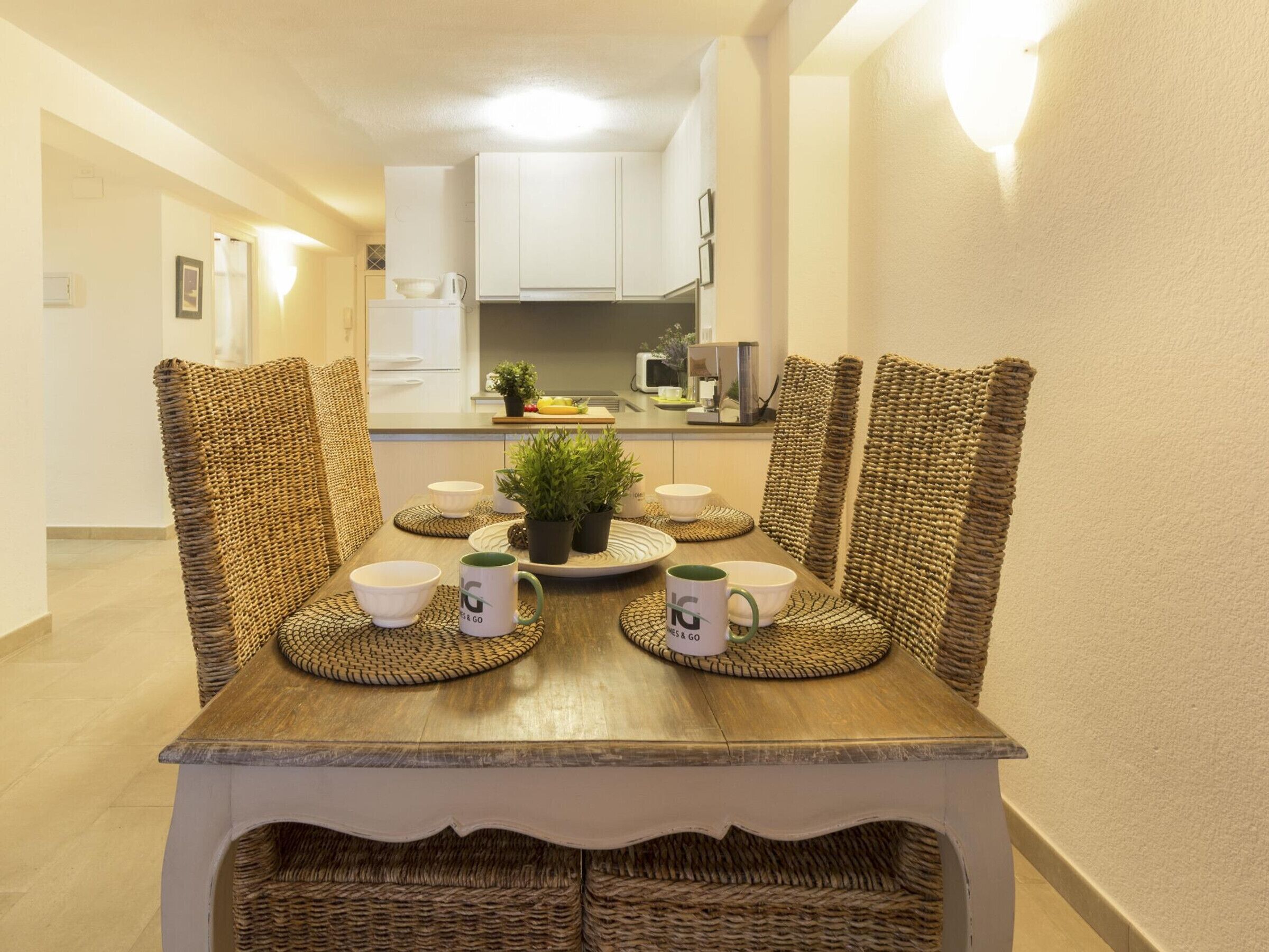 A dining table with woven chairs, a central plant, and a tray with bowls and mugs.