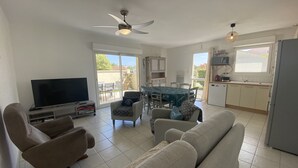 Sala de estar con sofá, sillones, televisor y ventilador de techo. Hay un comedor con mesa y sillas, y una cocina con alacenas y refrigerador.