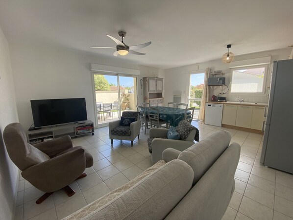 A living room with a sofa, armchairs, a TV, and a ceiling fan. There is a dining area with a table and chairs, and a kitchen with cabinets and a refrigerator. - Peaceful modern house in Fouras - 6 guests (Fouras)