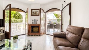 Un salón amplio con un sofá de cuero, una mesa de centro de vidrio y una chimenea. Hay un balcón con barandilla blanca y plantas en macetas.