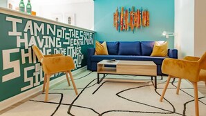 A modern living room with a blue sofa, two yellow chairs, a wooden coffee table, and a green wall with a motivational quote.
