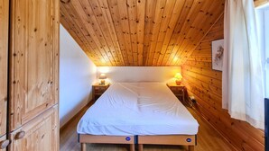 Ein kleiner, gemütlicher Raum mit Holzwänden und -decke, einem Einzelbett, zwei Nachttischen mit Lampen sowie einem Fenster mit Vorhängen.