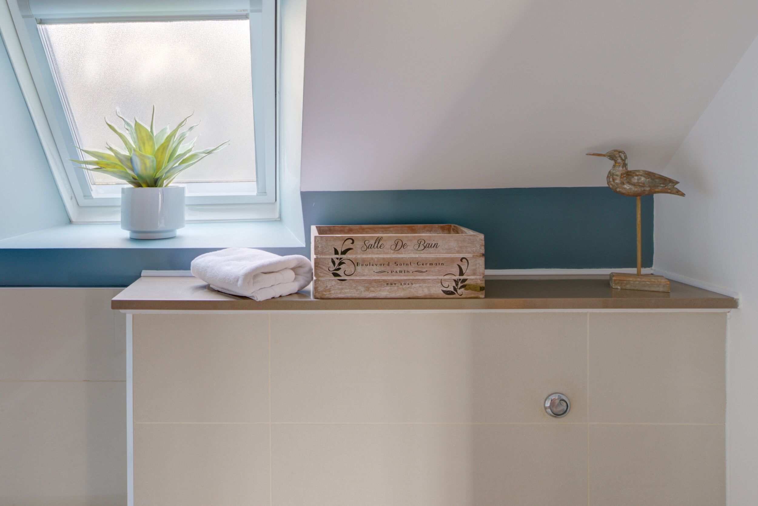 A bathroom with a window, a potted plant, a wooden box, a towel, and a bird figurine.