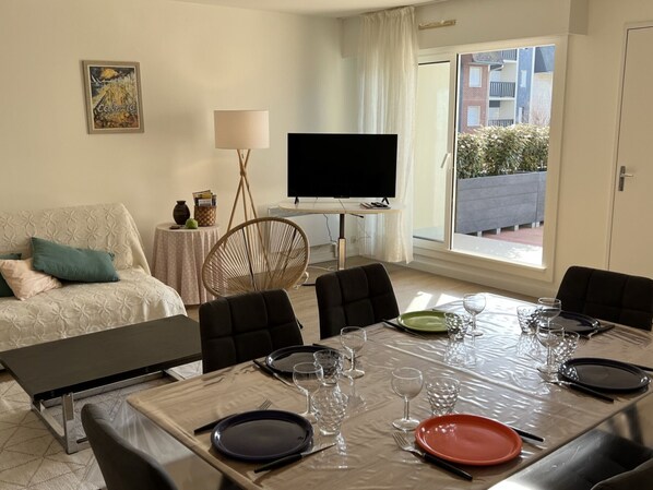 Un comedor con una mesa dispuesta para seis personas, un sofá, un televisor y una ventana con vistas a edificios.