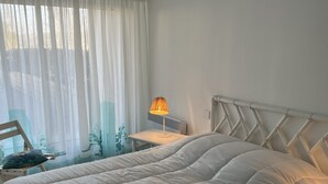 Una cama bien hecha con edredón blanco, una mesita de noche con lámpara y una silla con cojín.