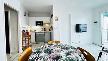 Une cuisine compacte avec des meubles blancs, un four à micro-ondes et un lave-vaisselle. On y trouve également un coin repas avec une table et des chaises, ainsi qu’un téléviseur fixé au mur.