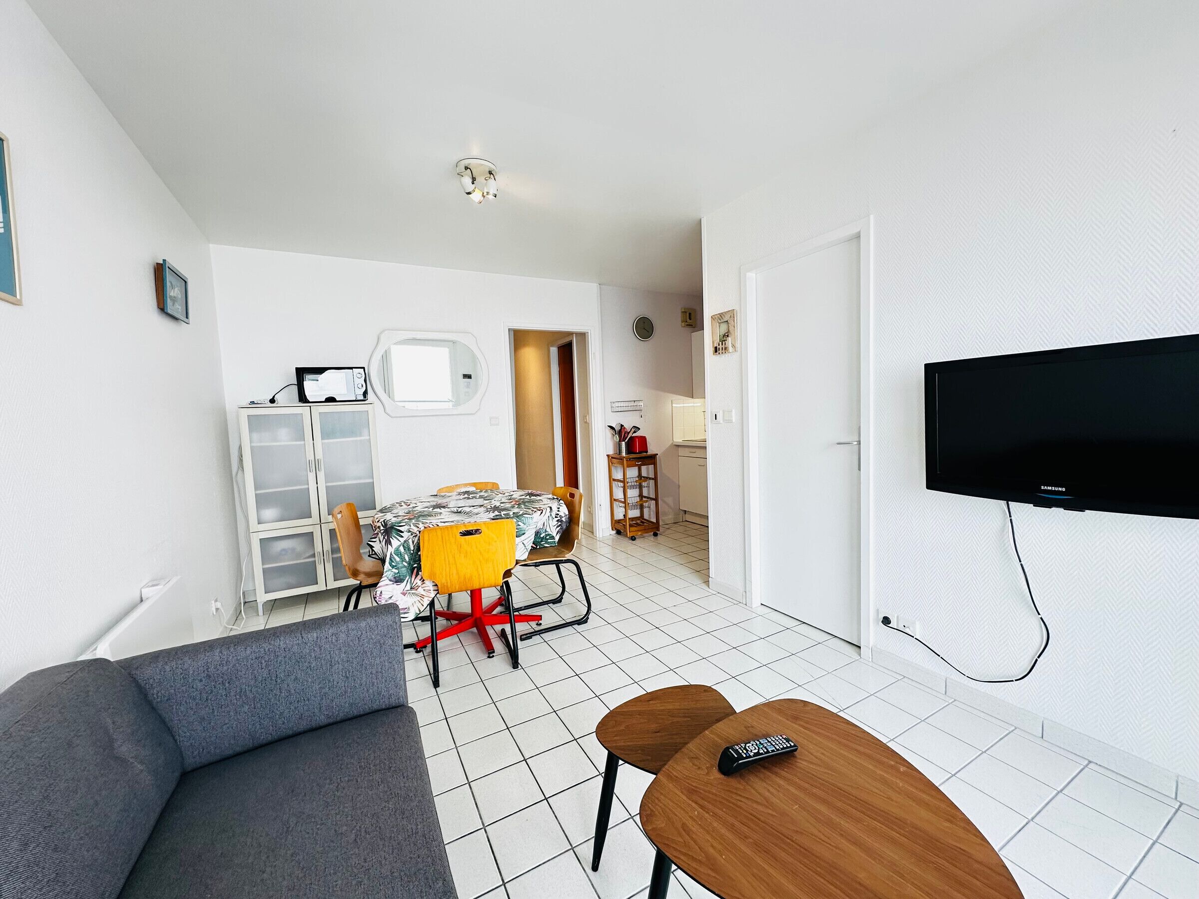 Un salon moderne avec un canapé gris, une table basse en bois, un téléviseur fixé au mur et un coin repas comprenant une table et des chaises.