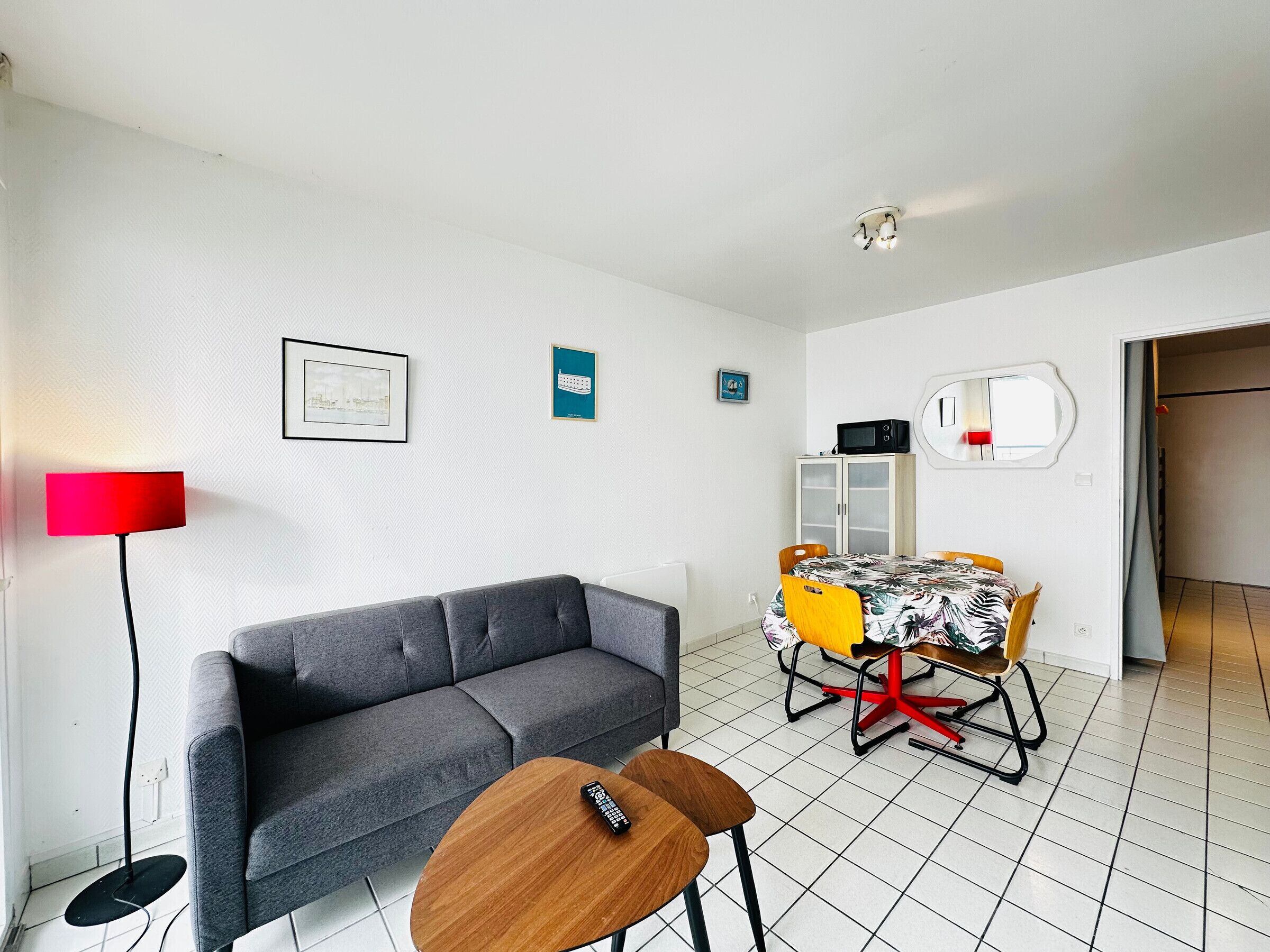A modern living room with a grey sofa, a dining table with chairs, a red floor lamp, and a ceiling light fixture.