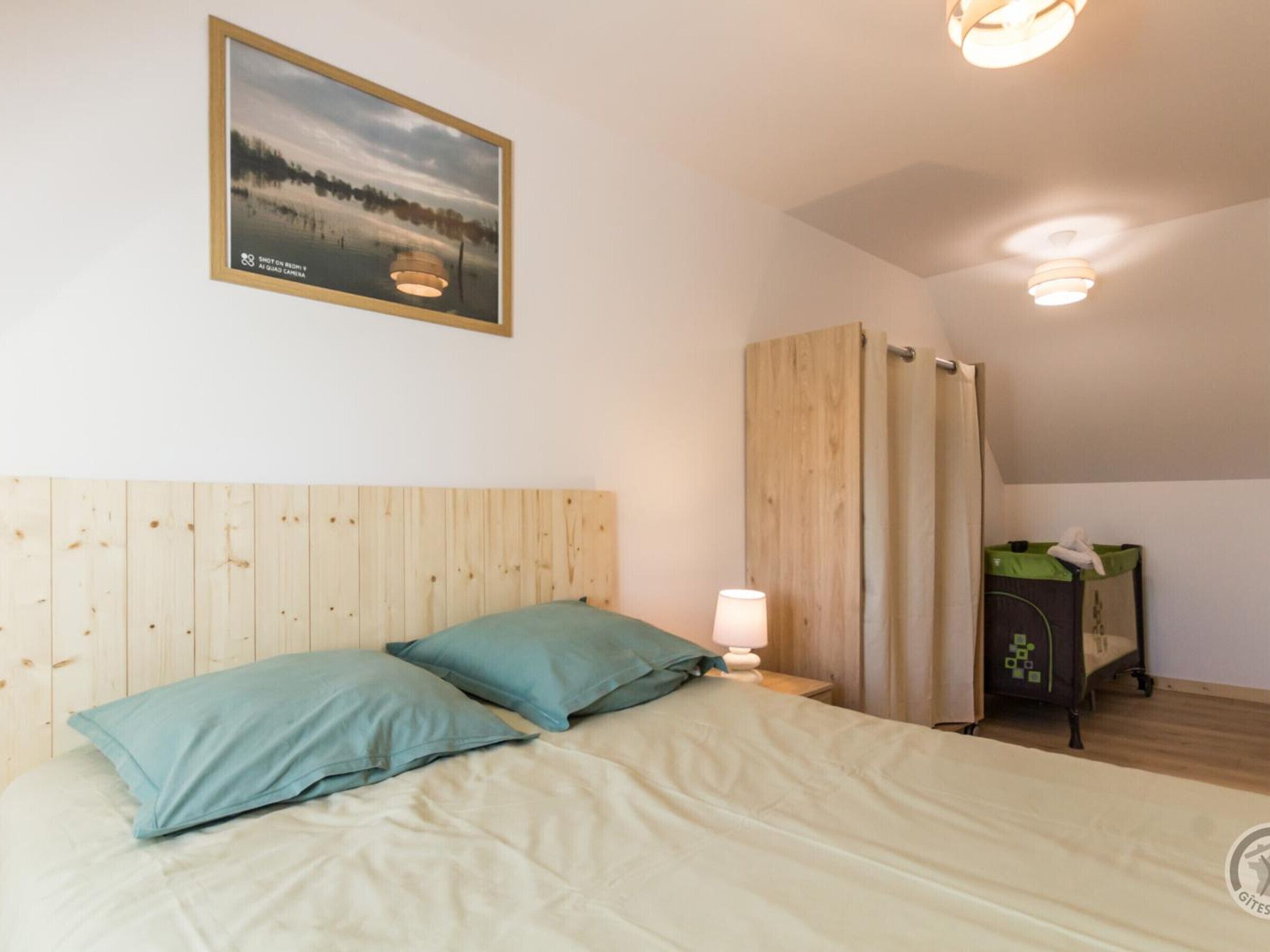 Ein Schlafzimmer mit Holzkopfstück, einem Bett mit zwei Kissen, einem Nachttisch mit Lampe, einem Fenster mit Vorhängen und einem kleinen Schrank mit grüner Ablagefläche.