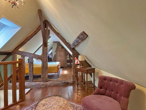 A loft bedroom with a sloped ceiling, wooden beams, a bed, a chair, and a small table.