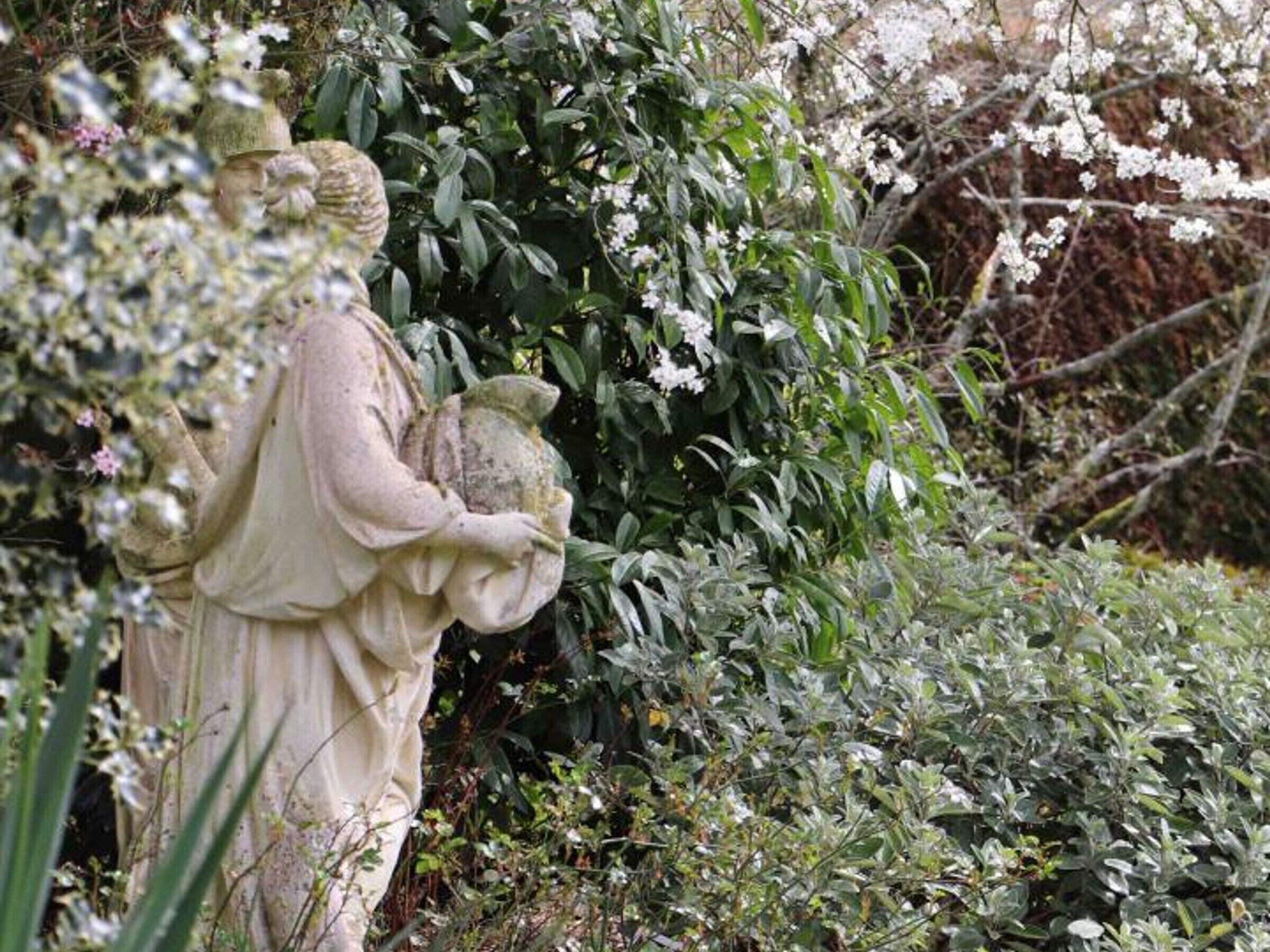Una estatua de una figura sosteniendo una piedra, rodeada de follaje denso y plantas con flores.