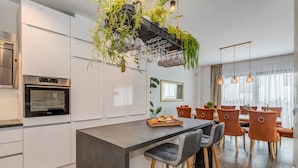 A modern kitchen with white cabinets, a built-in oven, a central island with bar stools, and a hanging plant arrangement.