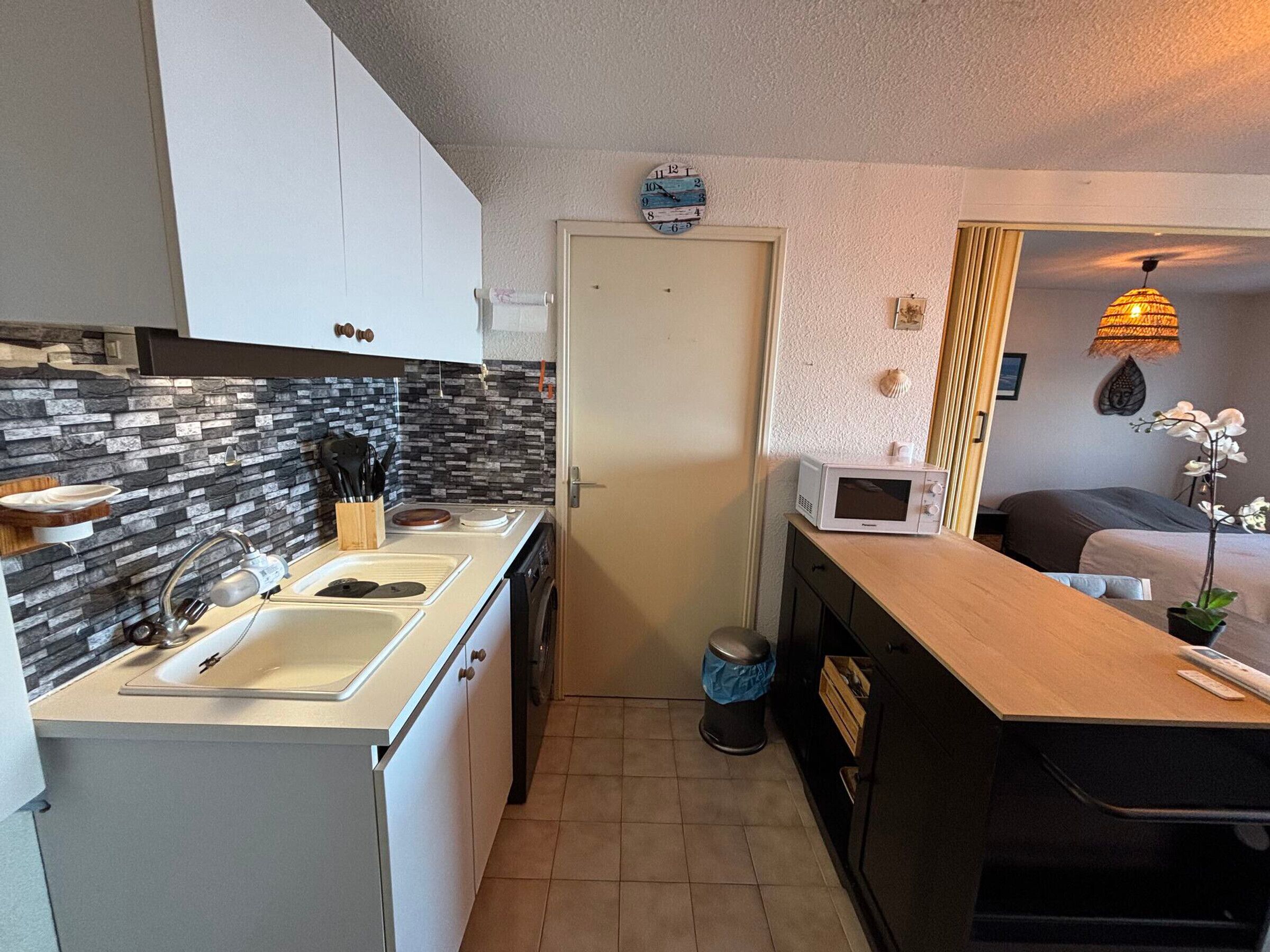 A compact kitchen with a sink, stove, and microwave. There's a door with a clock, a trash can, and a small plant on the counter.