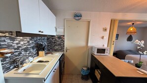 Cozinha compacta com pia, fogão e micro-ondas. Há uma porta com relógio, uma lixeira e uma pequena planta no balcão.