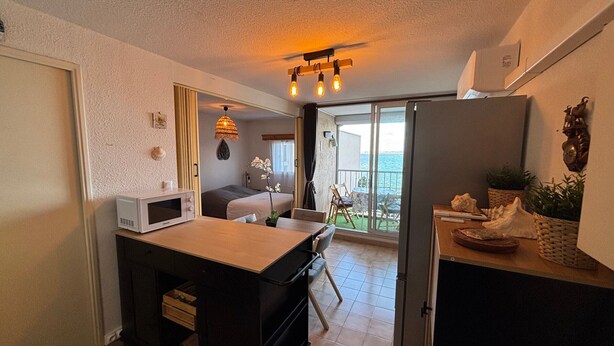 Cozinha compacta com micro-ondas, pia e geladeira. Área de refeições com mesa e cadeiras, e varanda com vista para o mar.