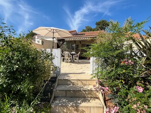 A pathway leading to a building with a covered patio area, outdoor seating, and a garden with flowering plants.