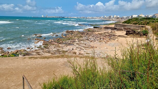 Eine Küstenlandschaft mit felsigem Gelände, einem Sandstrand und einer Stadtansicht in der Ferne.