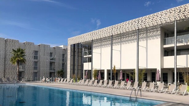 A swimming pool with lounge chairs and umbrellas, surrounded by a building with a patterned facade and balconies. - Stylish 2-Story Villa with Pool, Near Beach, Sleeps 6, Parking, A/C (Cap d'Agde)