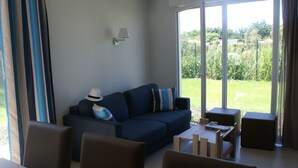 A modern living room with a dark blue sofa, a dining table with chairs, and a view of a garden through large glass doors. - Stylish 2-Story Villa with Pool, Near Beach, Sleeps 6, Parking, A/C (Cap d'Agde)