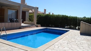Piscina com deck de ladrilho de pedra, área de estar coberta com cadeiras e um hedge ao fundo.