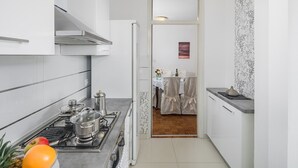 Een smalle keuken met een fornuis, pannen en een koffiezetapparaat. Op de achtergrond is een eethoek met een tafel en stoelen te zien.