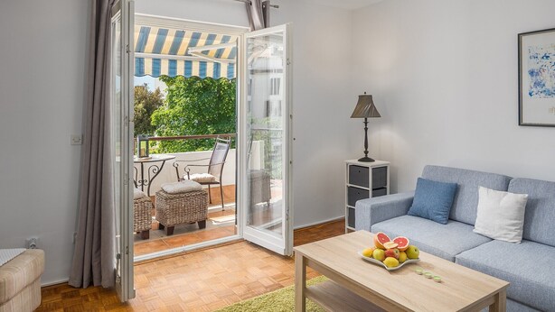 Een woonkamer met een bank, een bijzettafeltje met fruit, een lamp en een balkon met tuinmeubilair.