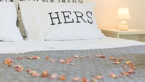 Een bed met witte kussens, waarvan één het woord 'HERS' heeft, en een hartvormige bloemblaarranrangement op de spreide.