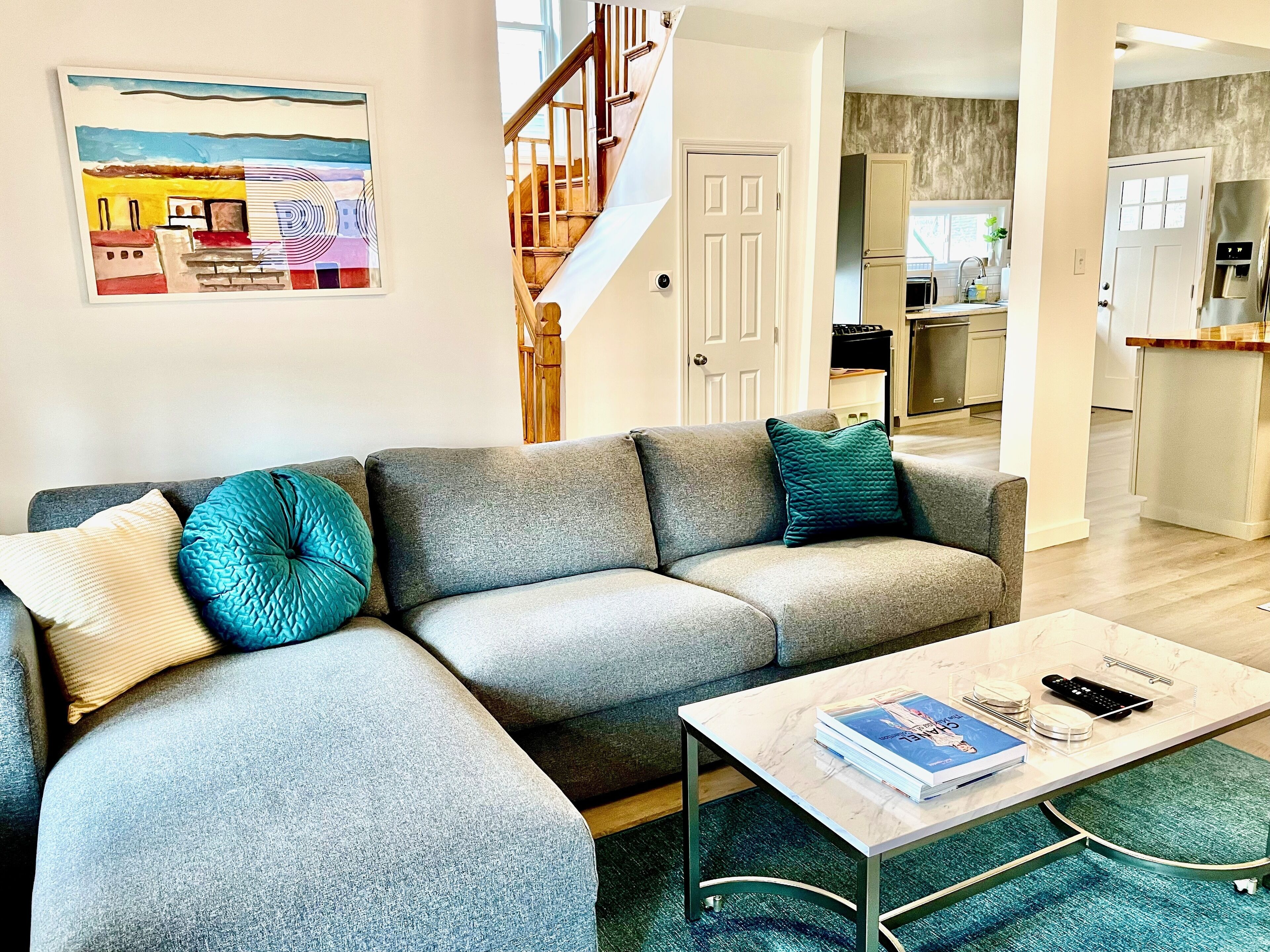 Un salon avec un canapé d’angle gris, des coussins turquoise, une table basse avec des livres et une télécommande, et une cuisine en arrière-plan.