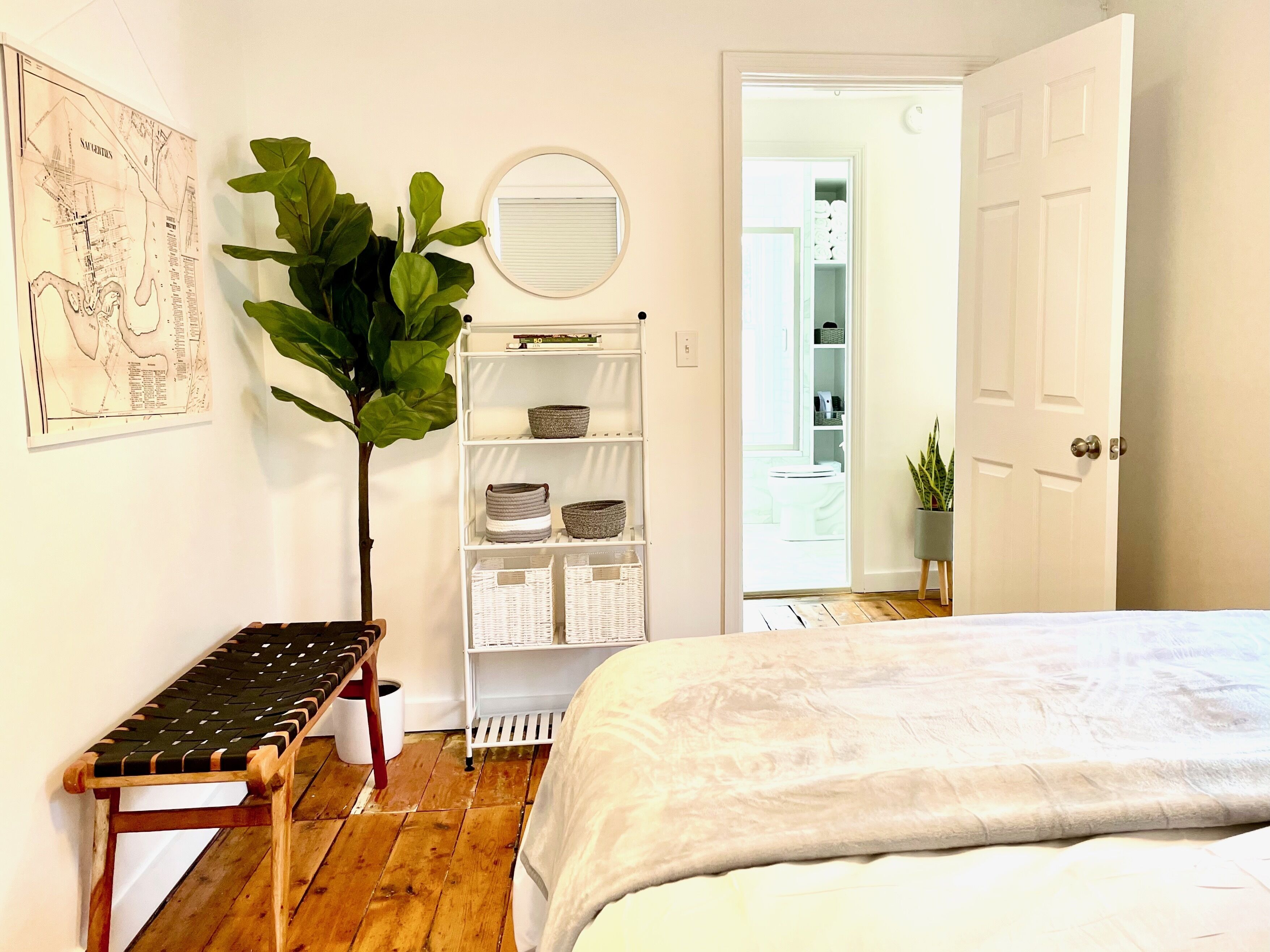 Une chambre à coucher comprenant un lit, un banc, une grande plante, une armoire blanche et une porte donnant sur une autre pièce.