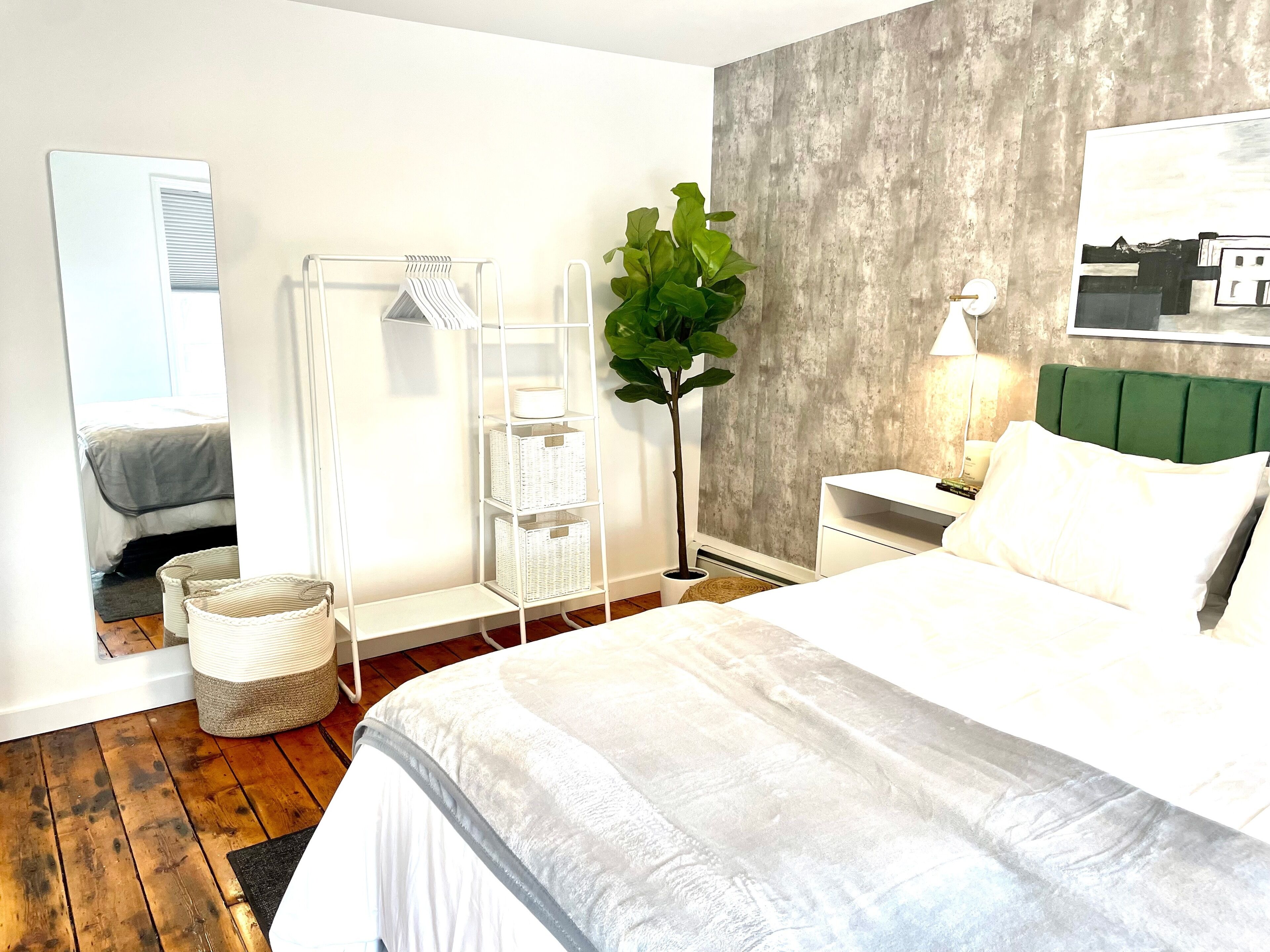 Une chambre d’hôtel moderne avec un grand lit, une table de chevet avec une lampe, une tête de lit recouverte d’un tissu vert, une étagère blanche, une plante en pot et un grand miroir.