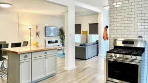A modern kitchen with a built-in stove, a dining area with a wooden table and chairs, and a living area with a sofa and a TV.