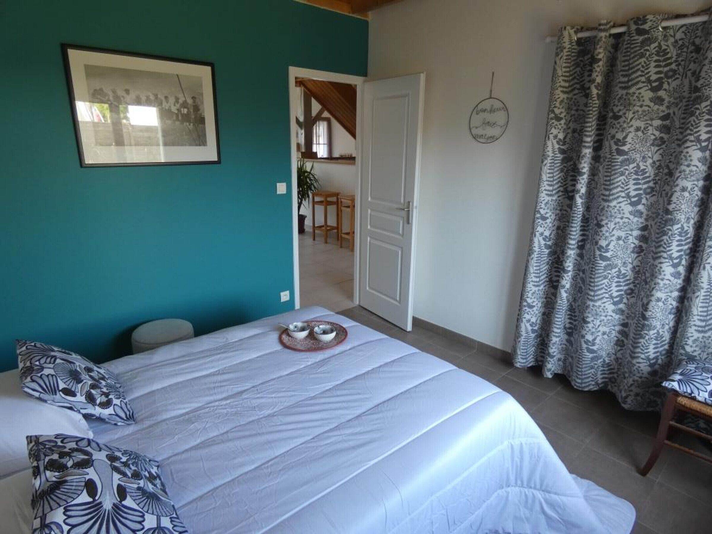 A bedroom with a bed, a framed picture, a door leading to a dining area, and a curtain with a floral pattern.