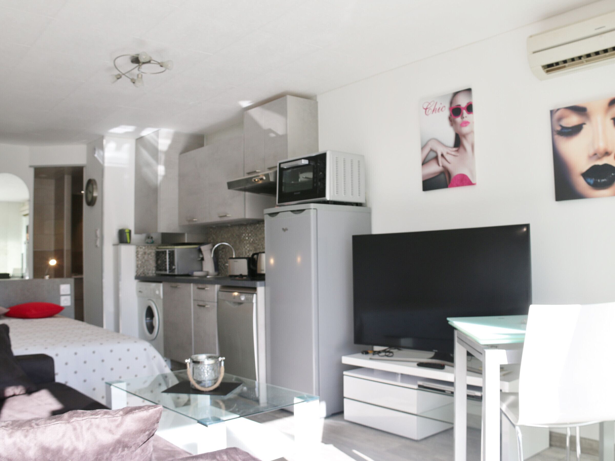 A modern, well-lit bedroom with a bed, bedside table, and a glass coffee table. Adjacent is a compact kitchenette with a microwave, kettle, and small refrigerator. A flat-screen TV is mounted on the wall, and there are two framed pictures above it.