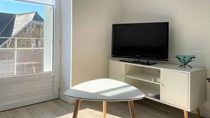 A room with a white TV stand, a flat-screen TV, a white table, and a window with blinds.