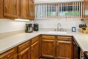 A kitchen with wooden cabinets, a white countertop, a coffee maker, a sink, and a window with blinds.